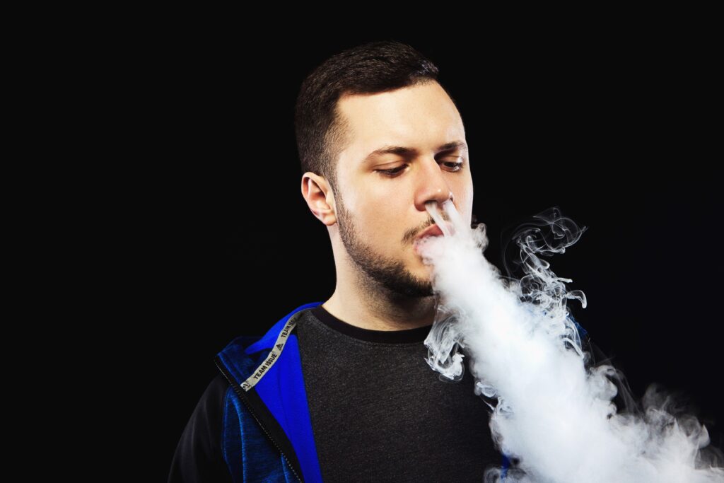 Mann in einer blauen Jacke, der eine große Dampfwolke ausatmet, während er eine Vape in der Hand hält, vor einem schwarzen Hintergrund.