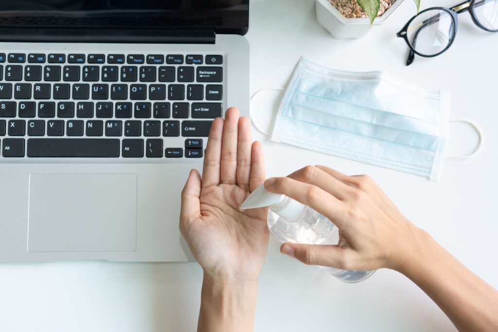 E-Learning Hygiene: Person desinfiziert Hände am Laptop-Arbeitsplatz mit Maske im Hintergrund