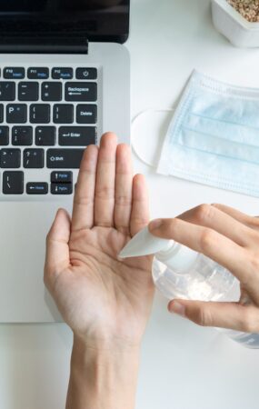 E-Learning Hygiene: Person desinfiziert Hände am Laptop-Arbeitsplatz mit Maske im Hintergrund
